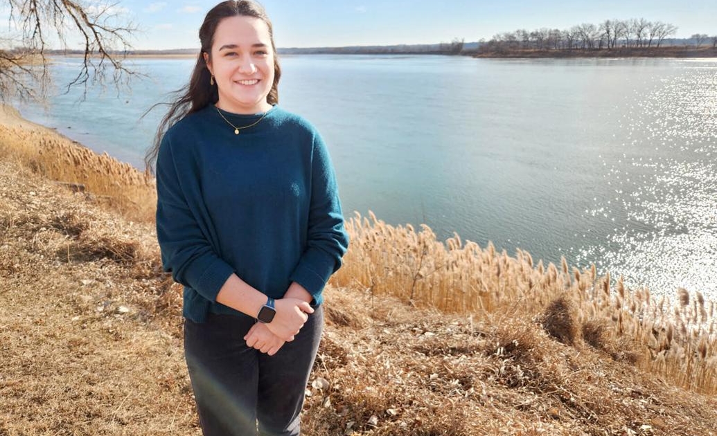 A woman stands along a grassy river bank with a wide smooth river behind her.