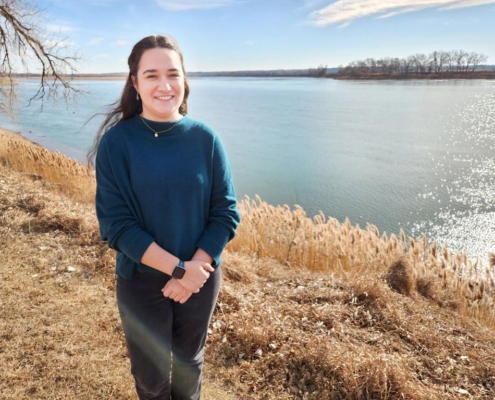 A woman stands along a grassy river bank with a wide smooth river behind her.
