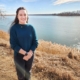 A woman stands along a grassy river bank with a wide smooth river behind her.