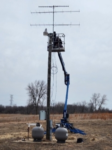 A tall metal tower with antennas across it gets installed in an open field