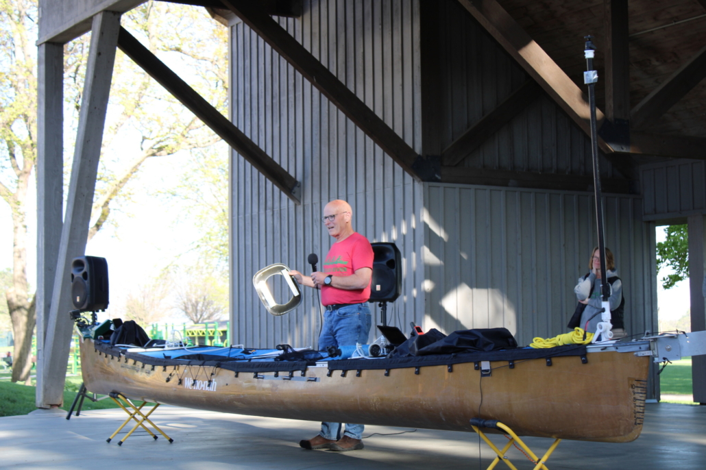 A man stands behind a canoe on a stage, holding a microphone and a piece of equipment