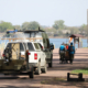 A van and truck trailering a boat follow behind a group of people wearing lifejackets