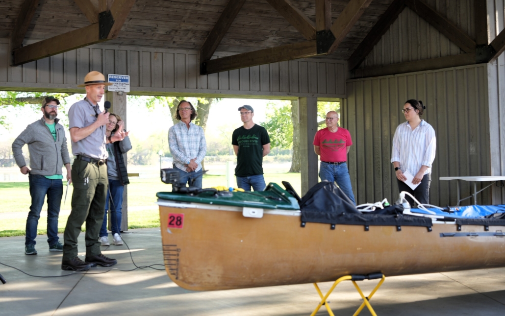 Seven people stand behind a canoe on a stage